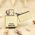 Gold Zippo lighter with its lid open, engraved 'On the way Route 66' on a sandy surface with seashells nearby.
