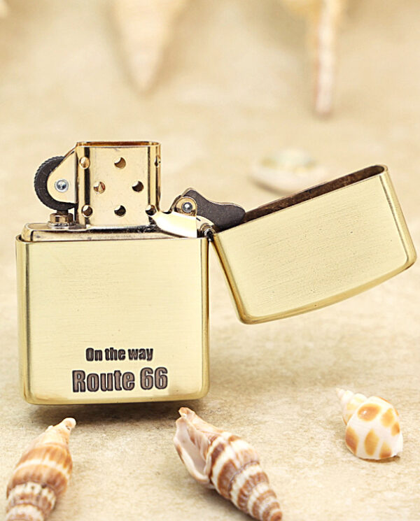 KZ160-03 Gold Zippo lighter with its lid open, engraved 'On the way Route 66' on a sandy surface with seashells nearby.