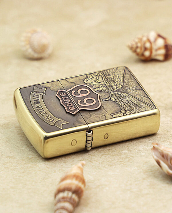 KZ160-04 Brass Zippo-style lighter with Route 66 emblem on a sandy surface, surrounded by seashells.