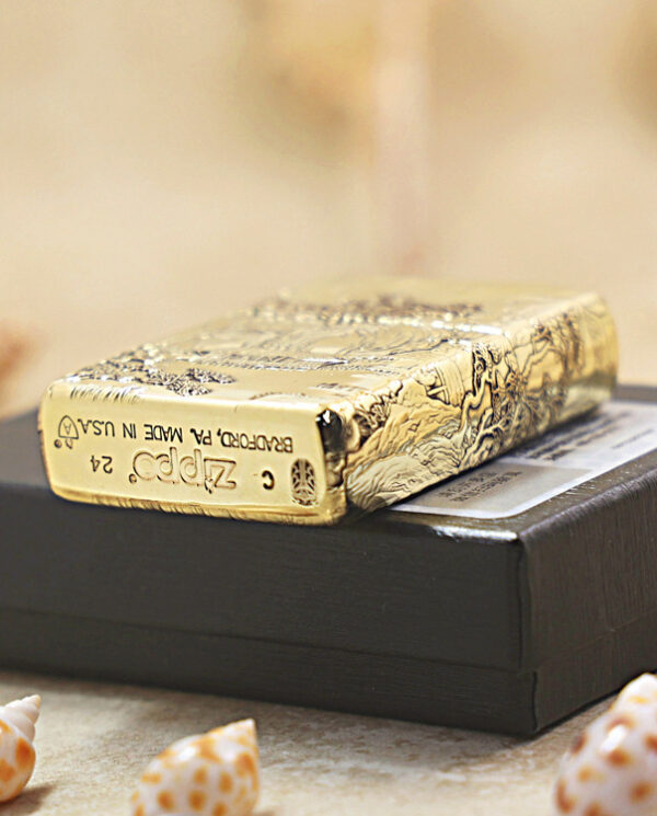 KZ162-05 Gold Zippo lighter with engraved pattern sits on a black box, with beach shells in the foreground.