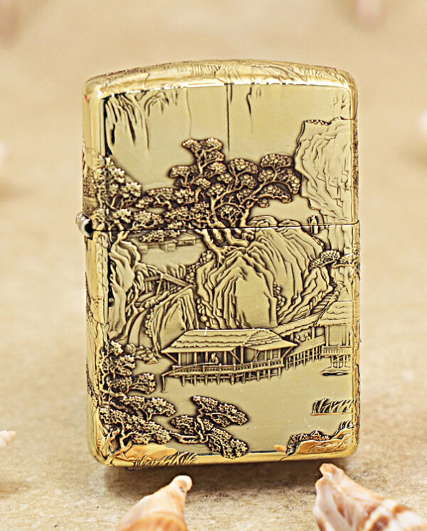 KZ162 Gold Zippo lighter with an intricate landscape engraving of trees, mountains, and a house, placed on sandy beach with seashells nearby