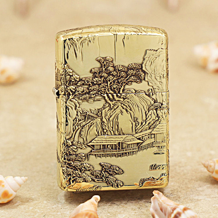 Gold Zippo lighter with an intricate landscape engraving of trees, mountains, and a house, placed on sandy beach with seashells nearby