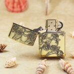 Gold lighter with ornate rose and vine engraving, opened to show the flame chamber on a sandy surface with seashells nearby.