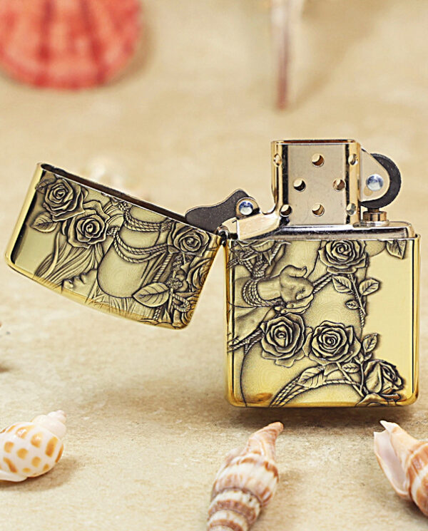 Gold lighter with ornate rose and vine engraving, opened to show the flame chamber on a sandy surface with seashells nearby.