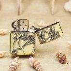 Gold lighter with black floral engraving, open lid, on sandy beach with seashells nearby.ang