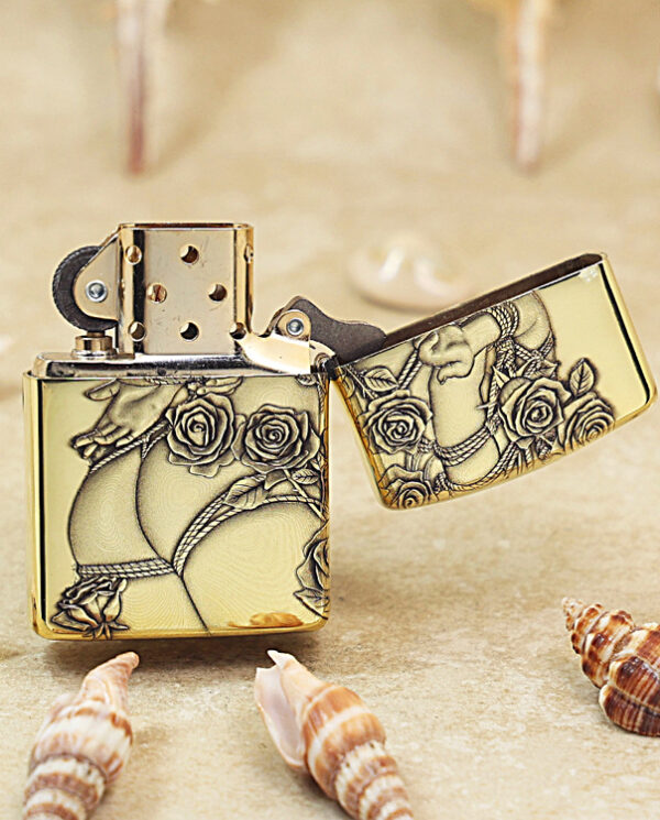 Gold lighter with black floral engraving, open lid, on sandy beach with seashells nearby.ang