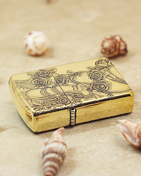 Gold engraved lighter with a rose motif rests on sandy beach among seashells.