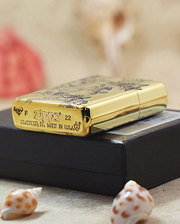 Gold Zippo lighter with floral engraving sits on a black box, surrounded by seashells on a light surface.