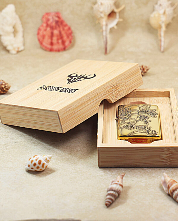 Gold Zippo-style lighter with a floral engraving in a wooden presentation box on a sandy surface with seashells around it.