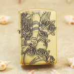 Gold-tone lighter with an embossed rose and vine design, resting on a sandy surface.
