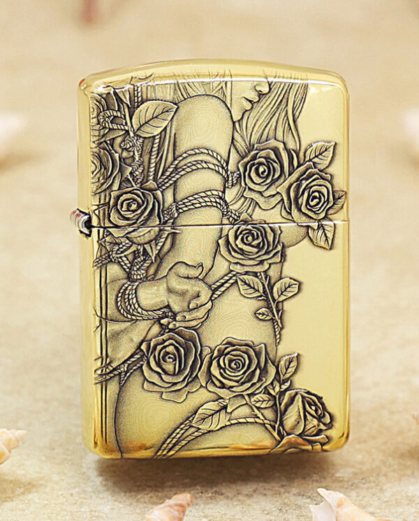 Gold-tone lighter with an embossed rose and vine design, resting on a sandy surface.