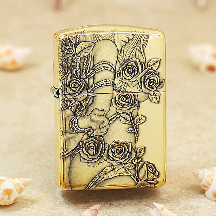 Gold-tone lighter with an embossed rose and vine design, resting on a sandy surface.