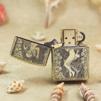 Ornate brass lighter with floral engravings and a partially open lid on a sandy beach with seashells nearby.