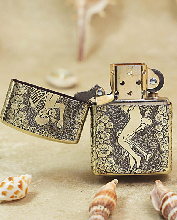 Ornate brass lighter with floral engravings and a partially open lid on a sandy beach with seashells nearby.