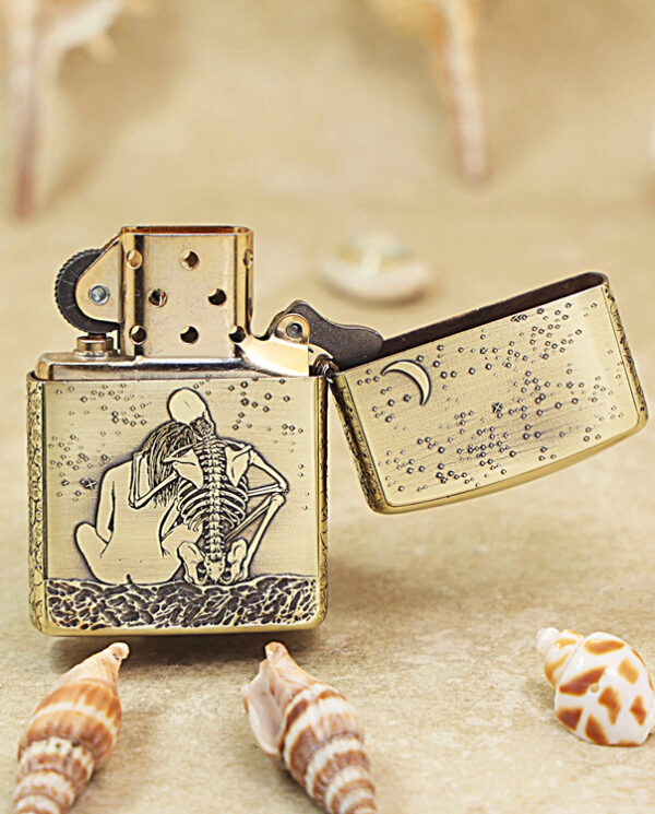 Gold Zippo-style lighter with a skeletal figure on the front, open lid, resting on a sandy surface with seashells nearby.