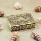 Gold-tone Zippo-style lighter with an engraved floral pattern and a reclining nude figure, resting on sand among seashells.