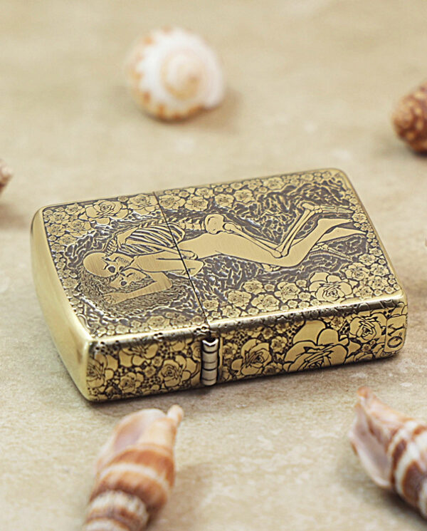 Gold-tone Zippo-style lighter with an engraved floral pattern and a reclining nude figure, resting on sand among seashells.