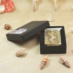Gold-toned Zippo lighter with an engraved man running design in an open black box on a sandy beach with seashells nearby.