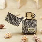 Snakeskin-pattern Zippo lighter with open lid on a sandy beach, surrounded by seashells and shells.
