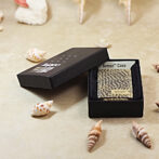 Square, snake-skin textured Zippo Armor Case inside an open black presentation box on a beige surface with scattered seashells nearby.