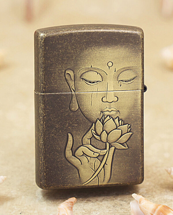 KZ179-01 Dark metal lighter etched with a meditating face and a hand holding a lotus; seashells in the background on a sandy surface.