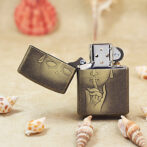 Brass Zippo lighter on sand with drawings: a finger to lips 'shh' motif and a sleepy face on the lid, surrounded by seashells on a beach