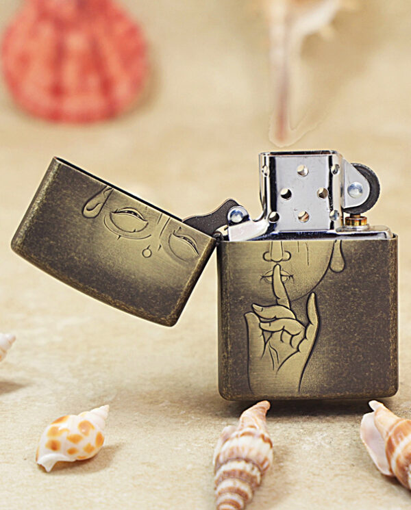 KZ179-02 Brass Zippo lighter on sand with drawings: a finger to lips 'shh' motif and a sleepy face on the lid, surrounded by seashells on a beach