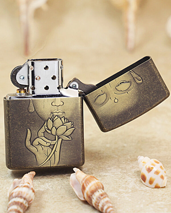 KZ179-03 Metal Zippo-style lighter with an engraved face and lotus flower, open on a sandy surface with seashells nearby.