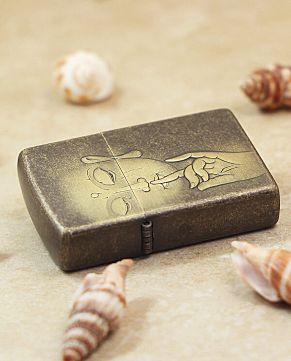KZ179-04 Metal lighter with an etched skull/face and hand motif, resting on sand among seashells on the beach.