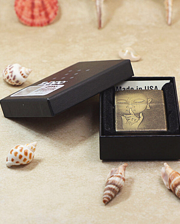 KZ179-06 Brass lighter with an engraved face inside a black box, the lid open and 'Made in USA' label visible among seashells.