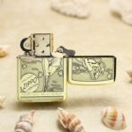 Gold Zippo lighter resting on sandy beach with seashells; lid open, flint wheel visible and engraved design on the body.