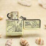 Gold Zippo lighter resting on sandy beach with seashells; lid open, flint wheel visible and engraved design on the body.