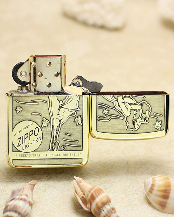 KZ183-03 Gold Zippo lighter resting on sandy beach with seashells; lid open, flint wheel visible and engraved design on the body.