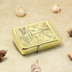 Gold Zippo lighter with engraved design lying on a sandy surface, surrounded by seashells.