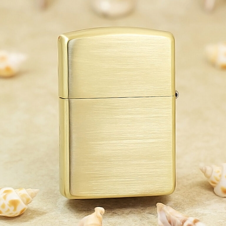 Brushed gold Zippo-style lighter standing upright on a sandy surface with scattered seashells nearby