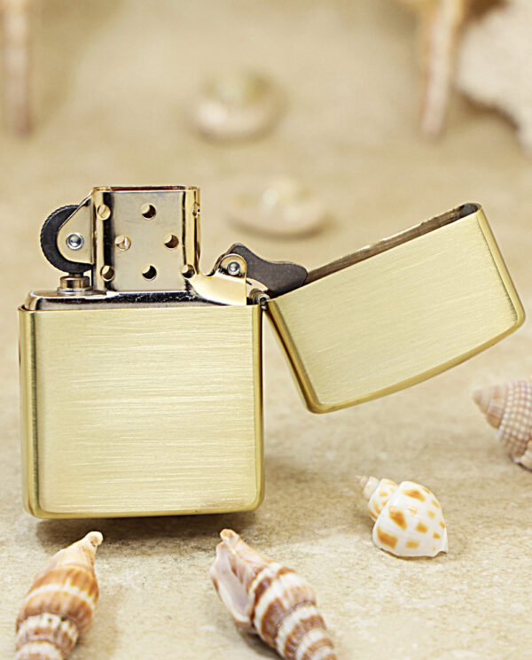 KZ184-03 Gold Zippo-style lighter with lid open, resting on sandy beach surrounded by seashells