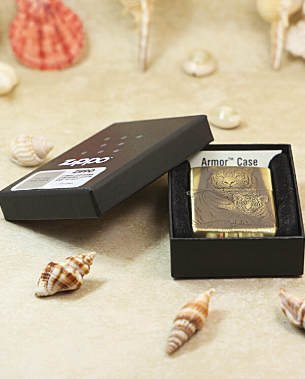 KZ184-06 Gold Zippo lighter in a black open box, labeled Armor Case, set on a sandy surface with seashells around it