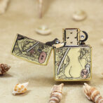Gold-tone Zippo lighter with intricate Asian-inspired engravings, lid open, resting on sand among seashells.