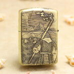 Gold Zippo-style lighter with a backward-facing samurai in armor, holding a spear, set against a mountainous village scene in relief on the surface.