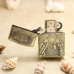 Brass lighter with an engraved medieval knight on the front, lid open, resting on sandy surface with seashells nearby.