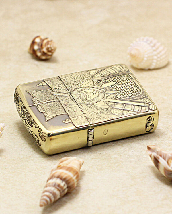 KZ190-04 Gold metal lighter with an engraved medieval scene resting on sandy beach among seashells.