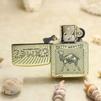 Gold-tone Zippo lighter with camel engraving and 'Nutty Menthol' banner, open on a sandy beach with seashells.