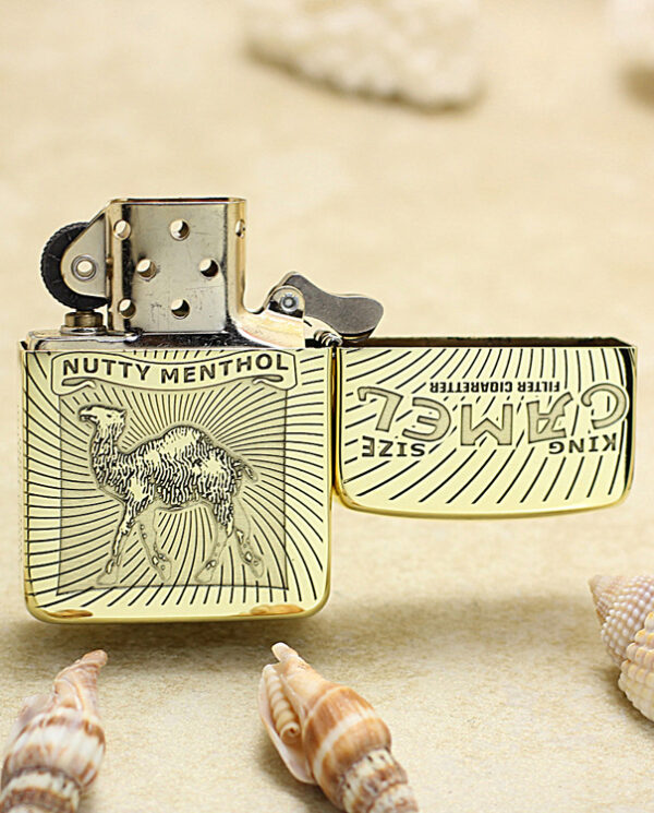 Gold-tone Zippo-style lighter with a camel engraving and 'Nutty Menthol' label, open lid on sandy beach among seashells.