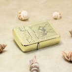 Gold-toned Zippo-style lighter on sandy surface with several seashells nearby, outdoors on the beach.