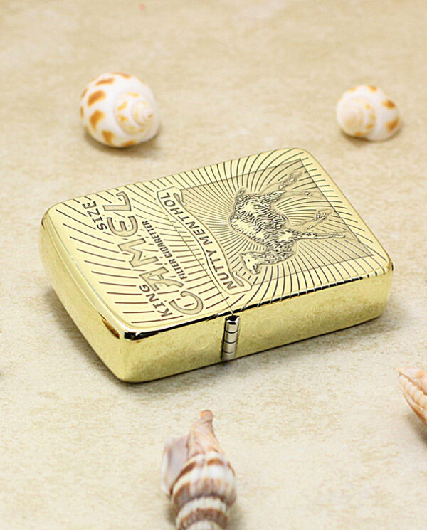 Gold-toned Zippo-style lighter on sandy surface with several seashells nearby, outdoors on the beach.