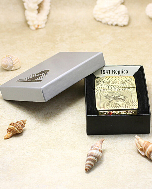 Gold 1941 Replica Camel cigarette tin inside a black box on a sandy surface, surrounded by seashells.