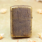 Brass Zippo-style lighter with a cracked leather texture standing upright on a sandy surface, surrounded by seashells.