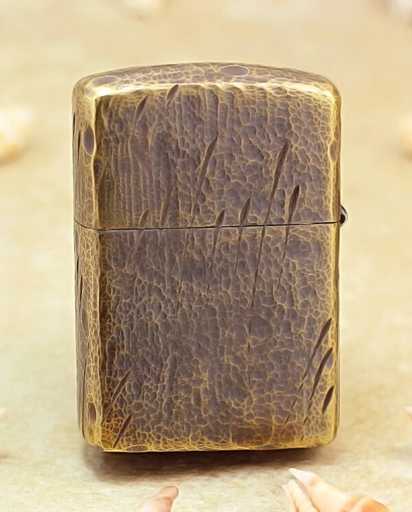 Brass Zippo-style lighter with a cracked leather texture standing upright on a sandy surface, surrounded by seashells.