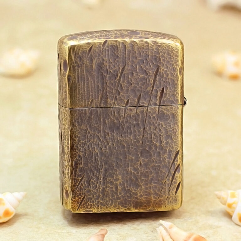 Brass Zippo-style lighter with a cracked leather texture standing upright on a sandy surface, surrounded by seashells.