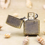 Vintage Zippo-style lighter with cracked patina on sandy surface, surrounded by seashells.